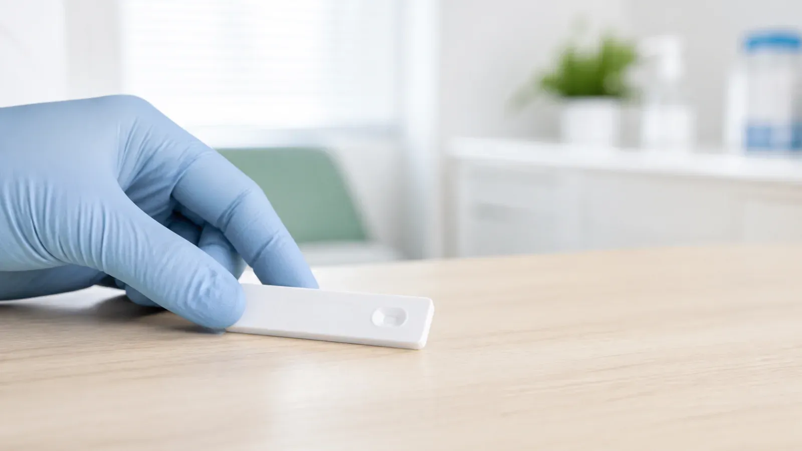 Dłoń w rękawiczce trzyma kasetkę szybkiego testu diagnostycznego na jasnym blacie.